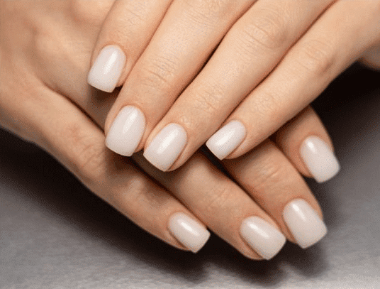 Hands with neatly manicured nails painted in a soft, neutral color rest on a gray surface.